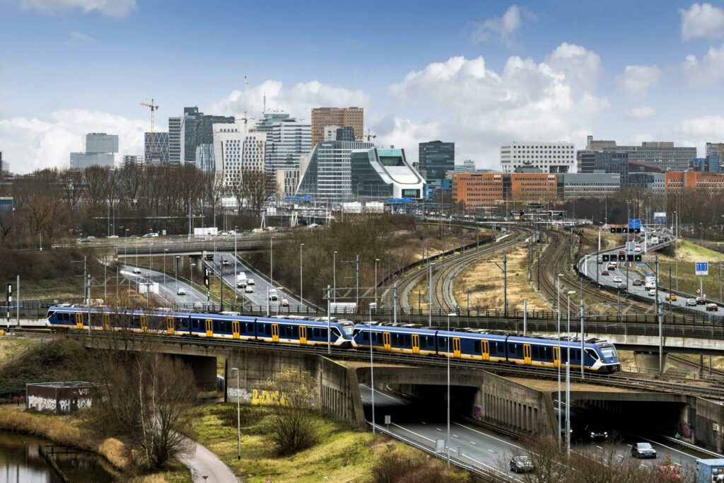 Een NS-trein rijdt over een viaduct bij de Johan Huizingalaan met op de achtergrond de moderne skyline van Amsterdam-Zuid.